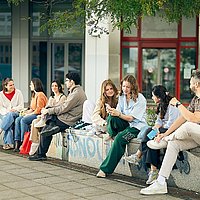 Gruppenfoto von Studierenden der ASH Berlin die vor der Hochschule sitzen und miteinander reden.