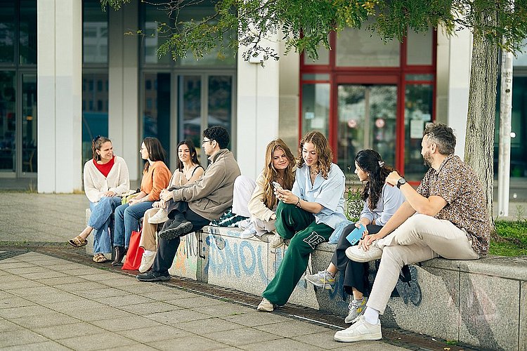 Gruppenfoto von Studierenden der ASH Berlin die vor der Hochschule sitzen und miteinander reden.