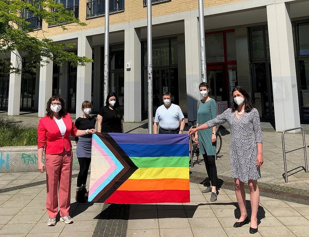 Bild der Regenbogenflaggenhissung davor stehen Teilnehmer mit dert Progress Flag