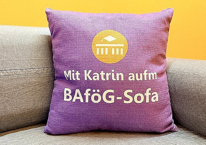 Ein lila Kissen mit der Aufschrift "Mit Katrin aufm BAFöG-Sofa" liegt auf einem grauen Sofa