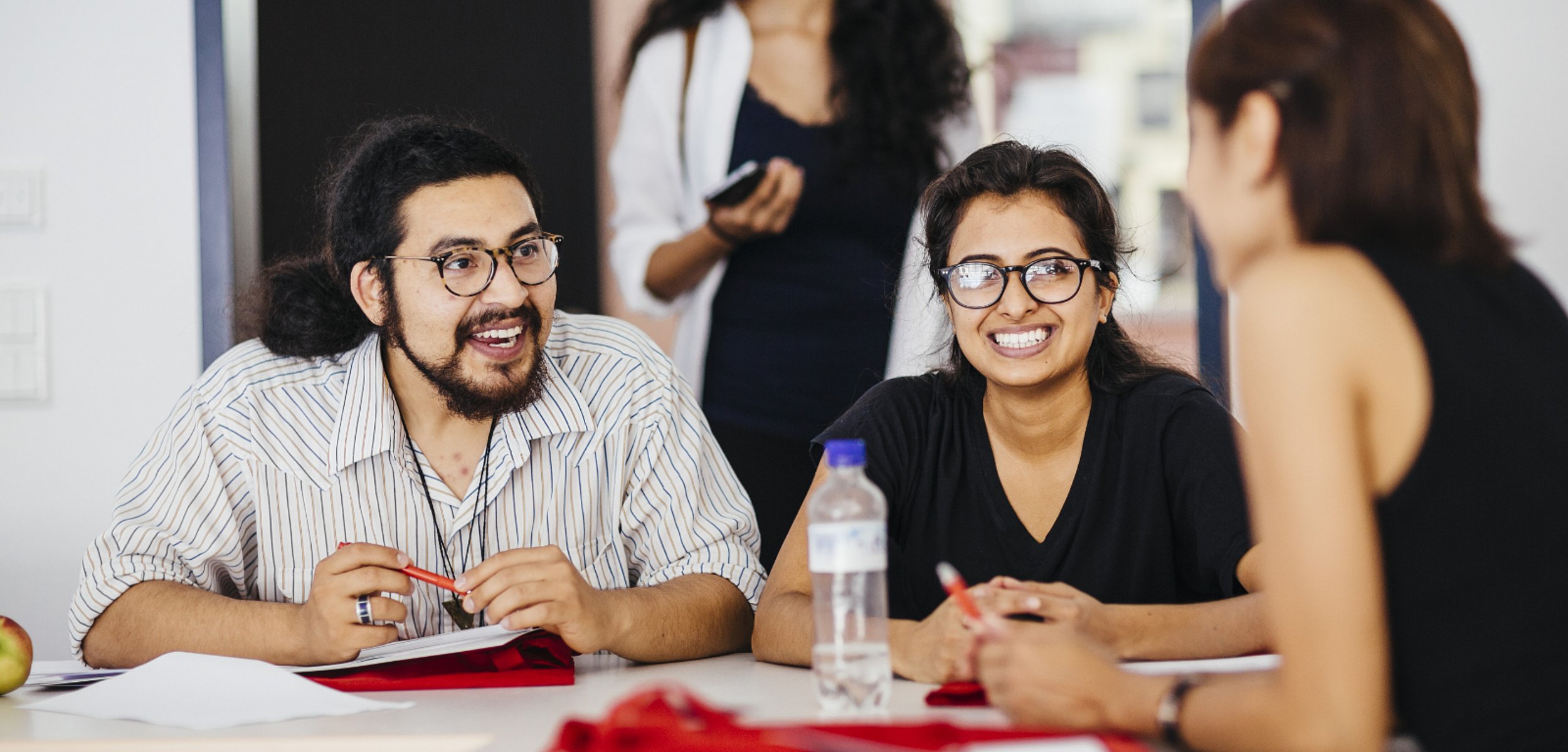 Eine Gruppe Studierender sitzt am Tisch, eine Studentin davon blickt lachend in die Kamera