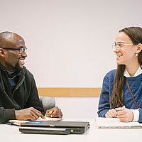 Auf dem Foto sind zwei Studierende zu sehen die nebeneinander an einem Tisch sitzen und sich unterhalten. 