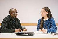 Auf dem Foto sind zwei Studierende zu sehen die nebeneinander an einem Tisch sitzen und sich unterhalten. 