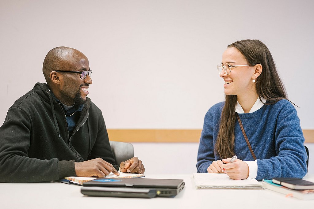 Auf dem Foto sind zwei Studierende zu sehen die nebeneinander an einem Tisch sitzen und sich unterhalten. 