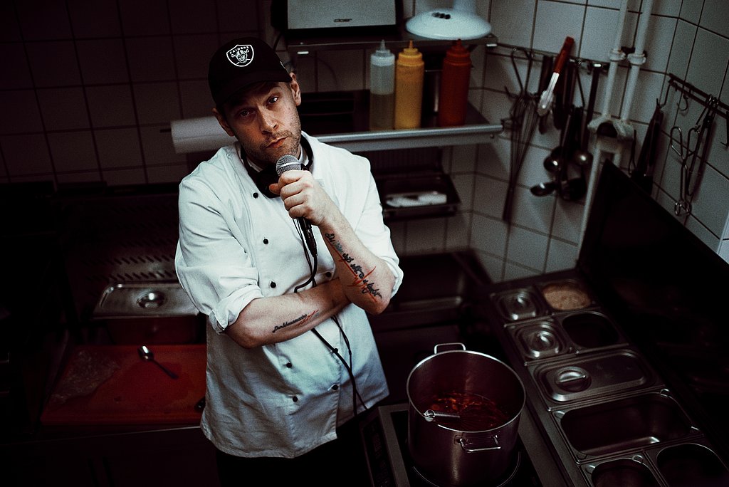 Paul Denkhaus mit weißer Kleidung in einer Küche neben einem Kochtopf und einem Mikrophon in der Hand