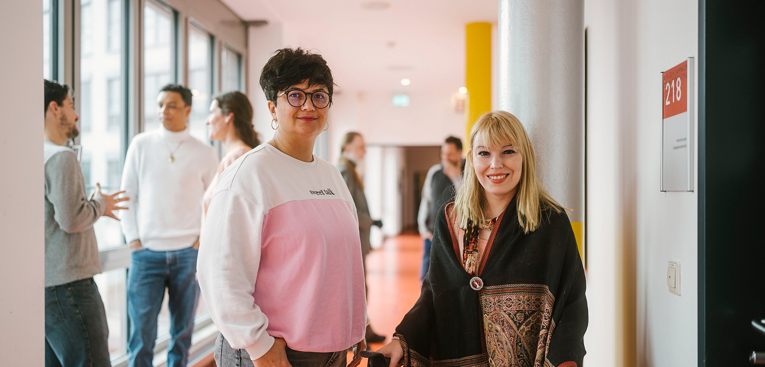 Studierende der ASH Berlin stehen im Flur der Hochschule und unterhalten sich. Zwei Personen lächeln in die Kamera.