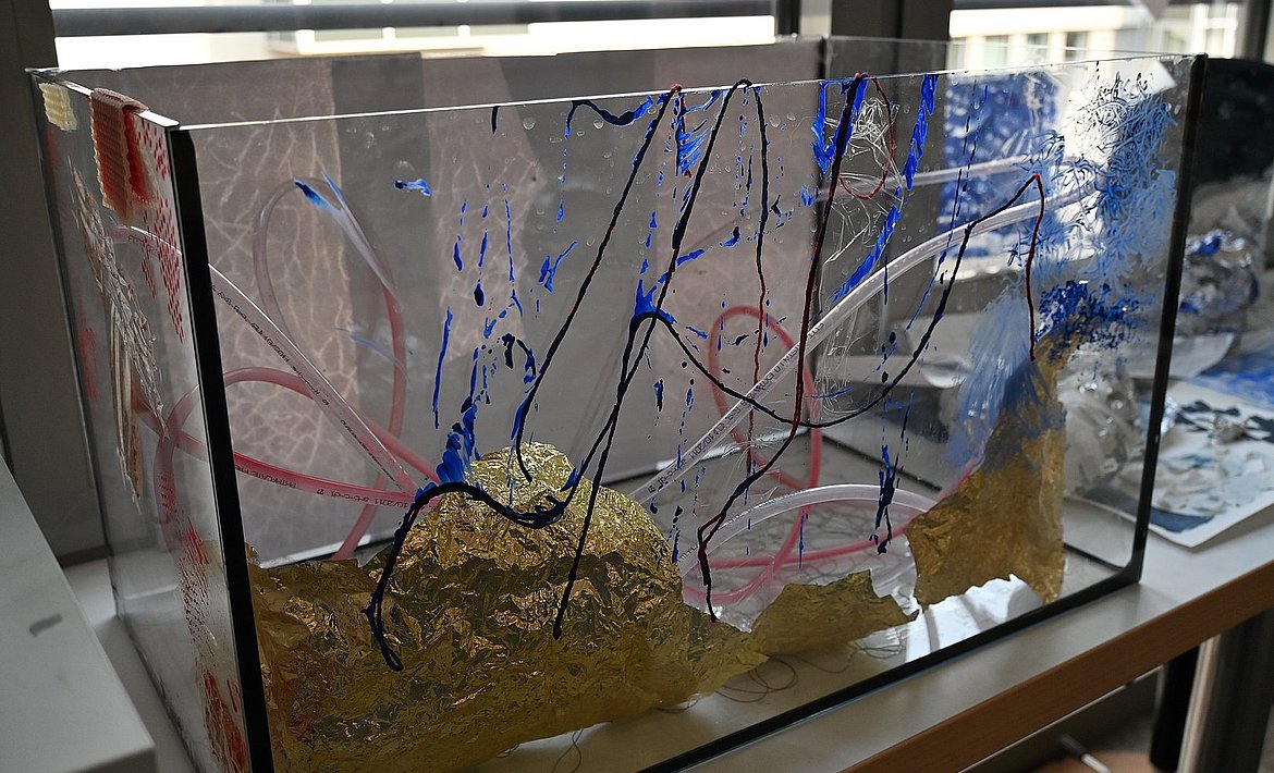 Es ist ein Glas-Körper zu sehen, in dem goldene Folie liegt und um diesen herum roter Faden in Kombination mit blauer Farbe gelegt wurde. 