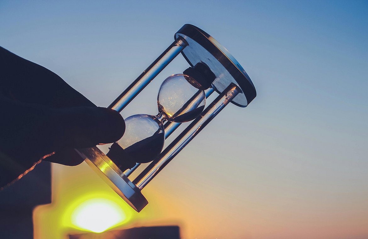 Eine Hand, die eine kleine Sanduhr vor dem Sonnenuntergang hält und die Kurzweiligkeit und Wichtigkeit des Moments symbolisiert.