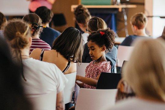 Kleines Mädchen sitzt in der Vorlesung neben ihrer Mutter und fragt sie etwas