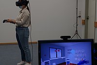 Auszubildende ausgestattet mit Virtual Reality-Brille und 2 Bewegungs-Controllern. Im Vordergrund steht ein Bildschirm auf dem ein virtuelles Krankenhauszimmer zu sehen ist.