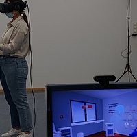 Auszubildende ausgestattet mit Virtual Reality-Brille und 2 Bewegungs-Controllern. Im Vordergrund steht ein Bildschirm auf dem ein virtuelles Krankenhauszimmer zu sehen ist.  