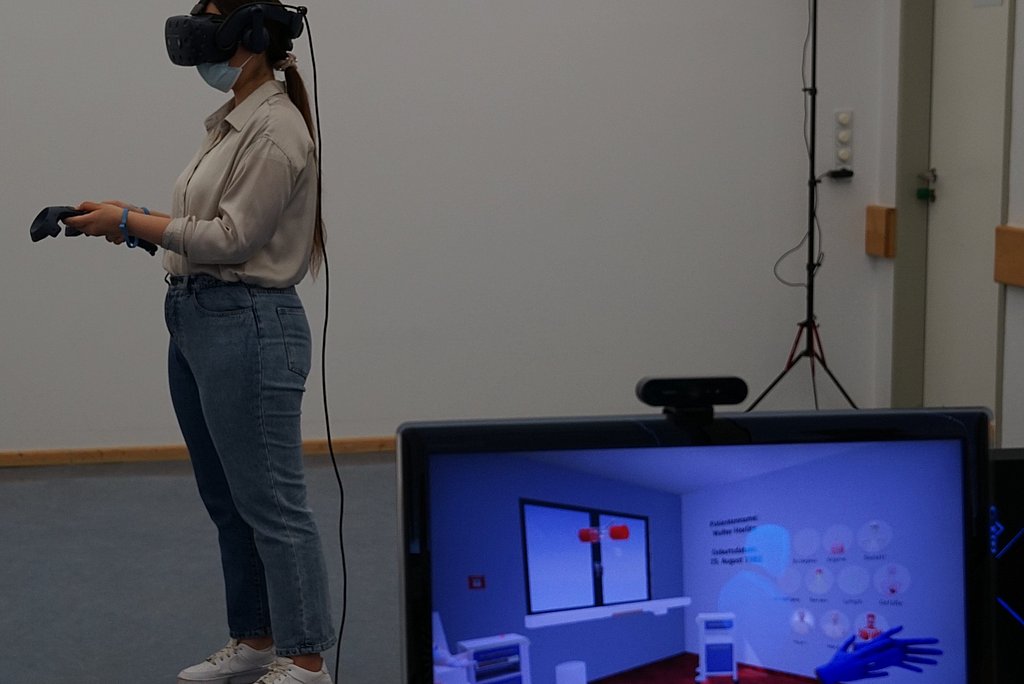 Auszubildende ausgestattet mit Virtual Reality-Brille und 2 Bewegungs-Controllern. Im Vordergrund steht ein Bildschirm auf dem ein virtuelles Krankenhauszimmer zu sehen ist.  