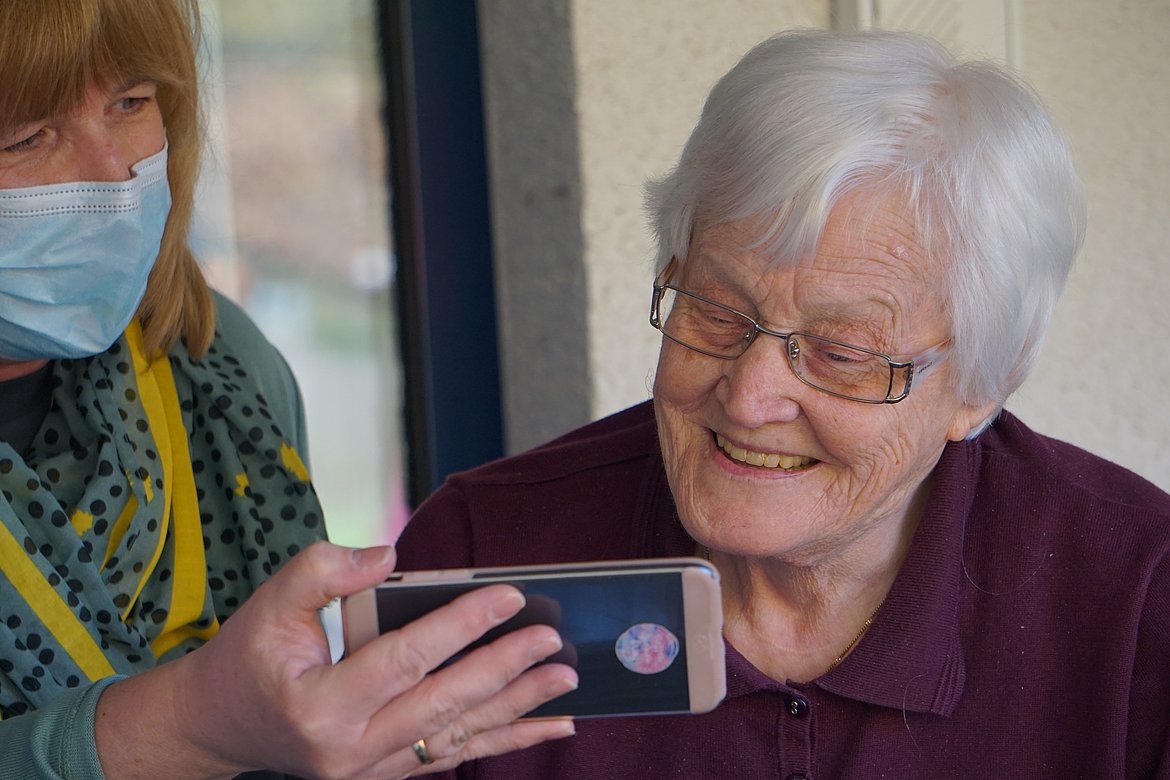 Eine ältere Frau schaut auf ein Handy, das von einer Pflegerin gehalten wird