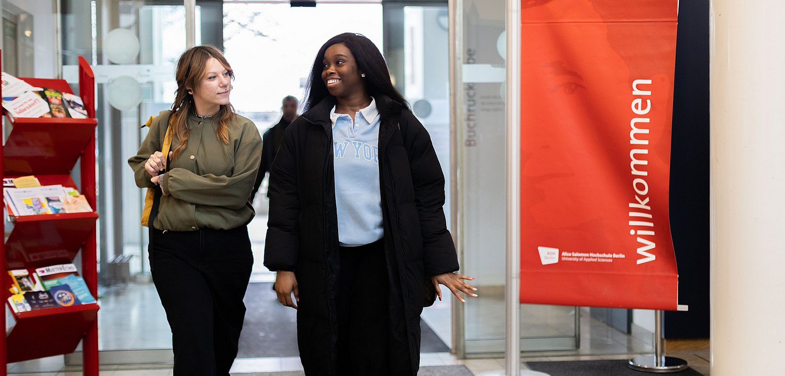 Zwei Studierende der ASH Berlin betreten die Hochschule und unterhalten sich. Rechts im Bild ist eine Fahne zu sehen, auf der das Wort Willkommen steht. 