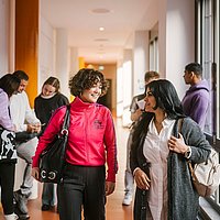 Auf dem Foto sind zwei ASH-Studierende zu sehen die einen Flur entlang laufen und sich dabei unterhalten. Im Hintergrund stehen weitere Personen, die sich ebenfalls unterhalten. 