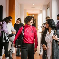 Auf dem Foto sind zwei ASH-Studierende zu sehen die einen Flur entlang laufen und sich dabei unterhalten. Im Hintergrund stehen weitere Personen, die sich ebenfalls unterhalten. 