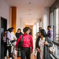 Auf dem Foto sind zwei ASH-Studierende zu sehen die einen Flur entlang laufen und sich dabei unterhalten. Im Hintergrund stehen weitere Personen, die sich ebenfalls unterhalten. 
