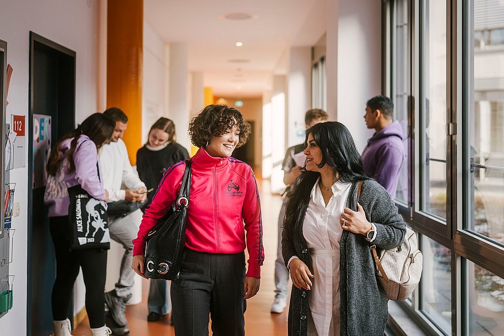 Auf dem Foto sind zwei ASH-Studierende zu sehen die einen Flur entlang laufen und sich dabei unterhalten. Im Hintergrund stehen weitere Personen, die sich ebenfalls unterhalten. 