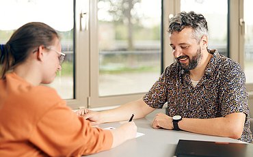 Zwei Studierende sitzen sich im Seminarraum gegenüber und sprechen miteinander.
