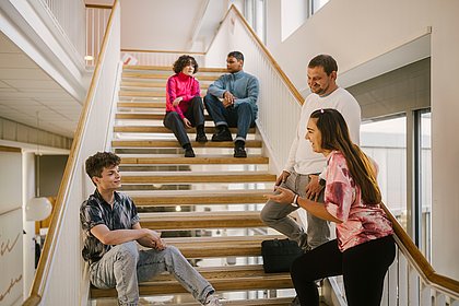 Auf dem Foto sind Studierende der ASH Berlin zu sehen, die auf einer Treppe in der Hochschule stehen oder sitzen und sich miteinander unterhalten.