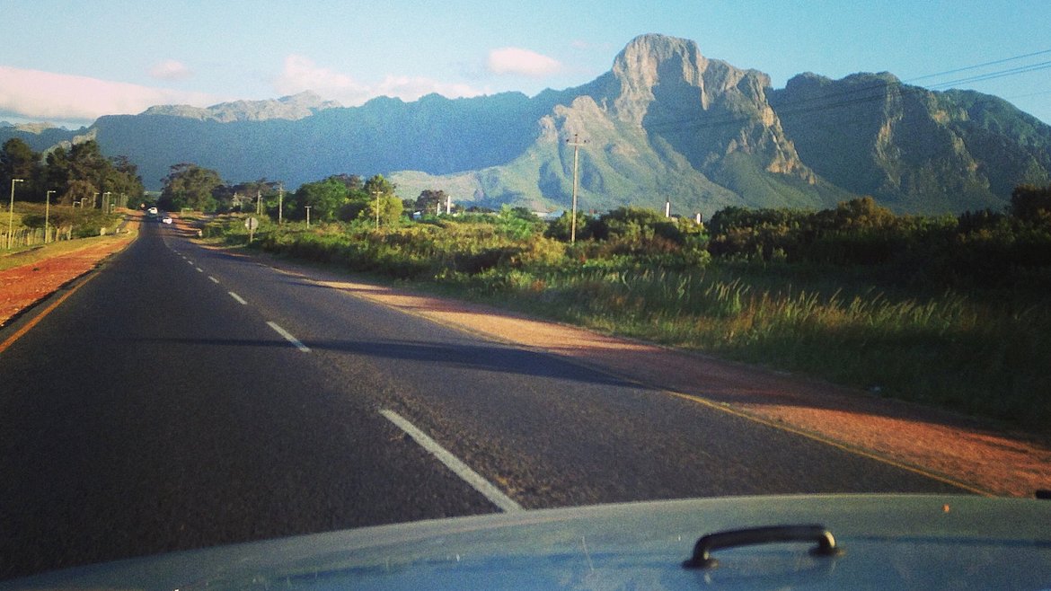 On the road_Südafrika: Aussicht aus einem Auto auf eine Straße mit Blick auf grünen Wegrand und Berge