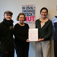 Auf dem Foto sind 4 Personen vor einer Wand zu sehen. Sie halten einen Bilderrahmen mit der Beitrittsurkunde in der Hand. Im Hintergrund ist ein Plakat zu sehen mit der Überschrift "In Zivilcourage steckt Mut"