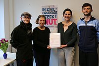 Auf dem Foto sind 4 Personen vor einer Wand zu sehen. Sie halten einen Bilderrahmen mit der Beitrittsurkunde in der Hand. Im Hintergrund ist ein Plakat zu sehen mit der Überschrift "In Zivilcourage steckt Mut"
