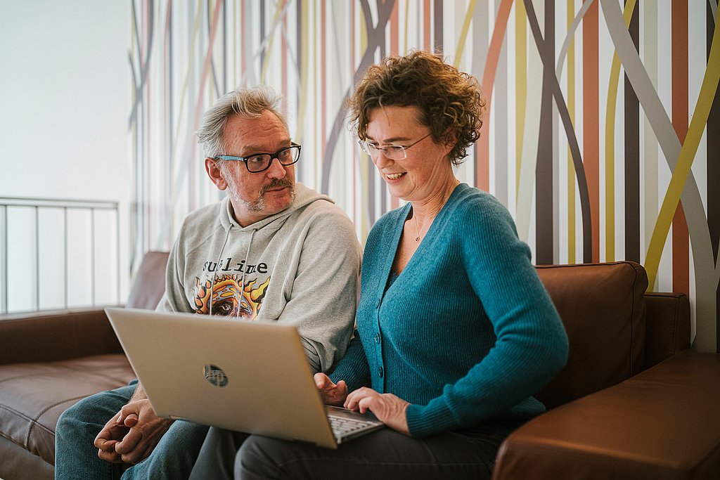 Zwei Studierende sitzen gemeinsam auf einer braunen Couch vor einem Laptop und sprechen miteinander. Im Hintergrund eine bunt gemusterte Wand.