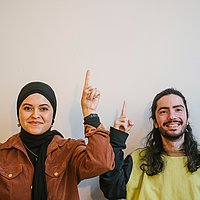 Zwei Studierende der ASH Berlin stehen vor einer weißen Wand und schauen lächelnd in die Kamera, dabei zeigen sie mit ihren Zeigefingern in Richtung Decke. 