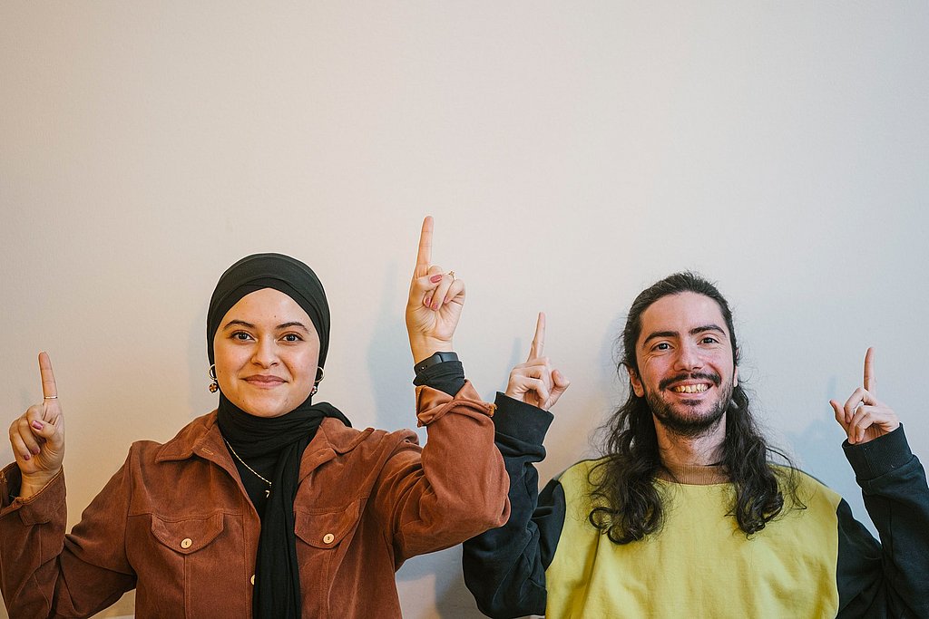 Zwei Studierende der ASH Berlin stehen vor einer weißen Wand und schauen lächelnd in die Kamera, dabei zeigen sie mit ihren Zeigefingern in Richtung Decke. 