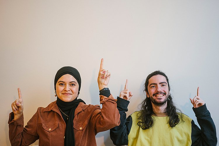 Zwei Studierende der ASH Berlin stehen vor einer weißen Wand und schauen lächelnd in die Kamera, dabei zeigen sie mit ihren Zeigefingern in Richtung Decke.