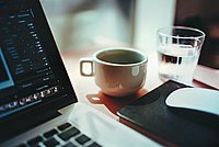 Laptop, Kaffeetasse und Wasserglas auf einem Holztisch.