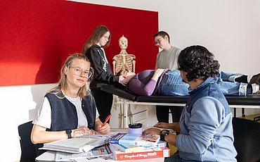 Zwei Studierende sitzen sich an einem Tisch mit vielen Büchern gegenüber. Ein Student schaut auf eine Gruppe von Studierenden hinter sich. Diese unterhalten sich gerade mit einer Studierenden, die auf einer Therapielliege liegt.