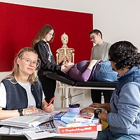 Zwei Studierende sitzen sich an einem Tisch mit vielen Büchern gegenüber. Ein Student schaut auf eine Gruppe von Studierenden hinter sich. Diese unterhalten sich gerade mit einer Studierenden, die auf einer Therapielliege liegt.