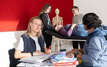 Zwei Studierende sitzen sich an einem Tisch mit vielen Büchern gegenüber. Ein Student schaut auf eine Gruppe von Studierenden hinter sich. Diese unterhalten sich gerade mit einer Studierenden, die auf einer Therapielliege liegt.
