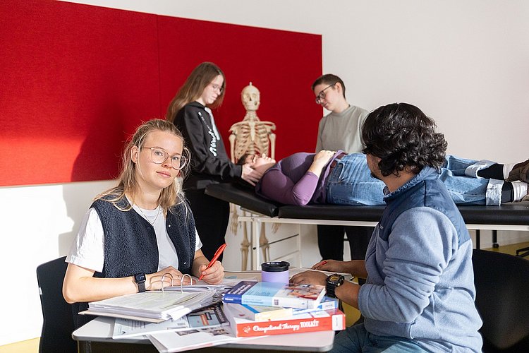 Zwei Studierende sitzen sich an einem Tisch mit vielen Büchern gegenüber. Ein Student schaut auf eine Gruppe von Studierenden hinter sich. Diese unterhalten sich gerade mit einer Studierenden, die auf einer Therapielliege liegt.