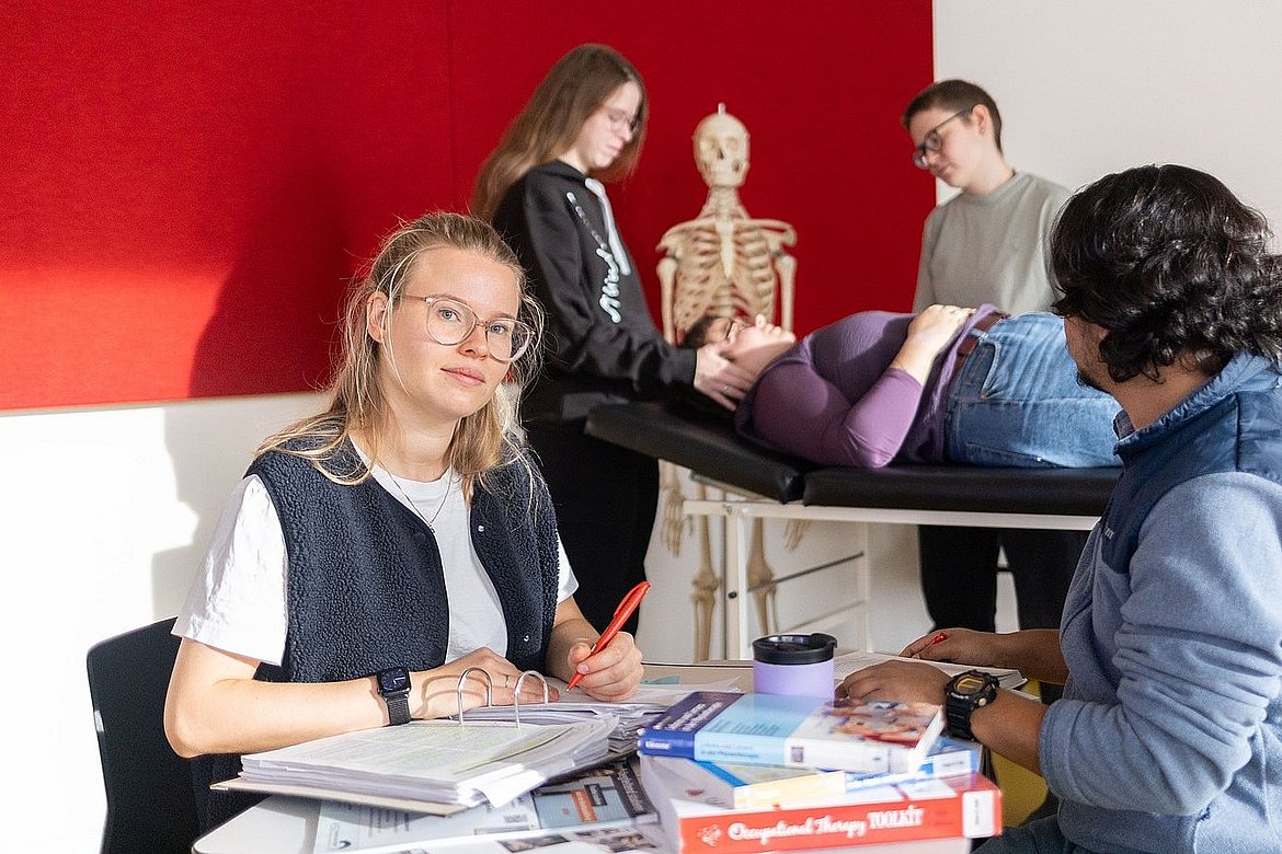 Zwei Studierende sitzen sich an einem Tisch mit vielen Büchern gegenüber. Ein Student schaut auf eine Gruppe von Studierenden hinter sich. Diese unterhalten sich gerade mit einer Studierenden, die auf einer Therapielliege liegt.