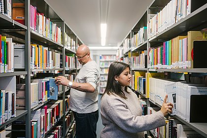 Zwei Personen in der Bibliothek, Bücher im Regal betrachtend.