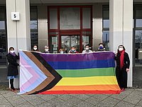 Die Kanzlerin der ASH Berlin und Mitarbeitende der Hochschule stehen vor der ASH Berlin und halten die Progressive Pride Flag hoch