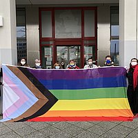 Die Kanzlerin der ASH Berlin und Mitarbeitende der Hochschule stehen vor der ASH Berlin und halten die Progressive Pride Flag hoch