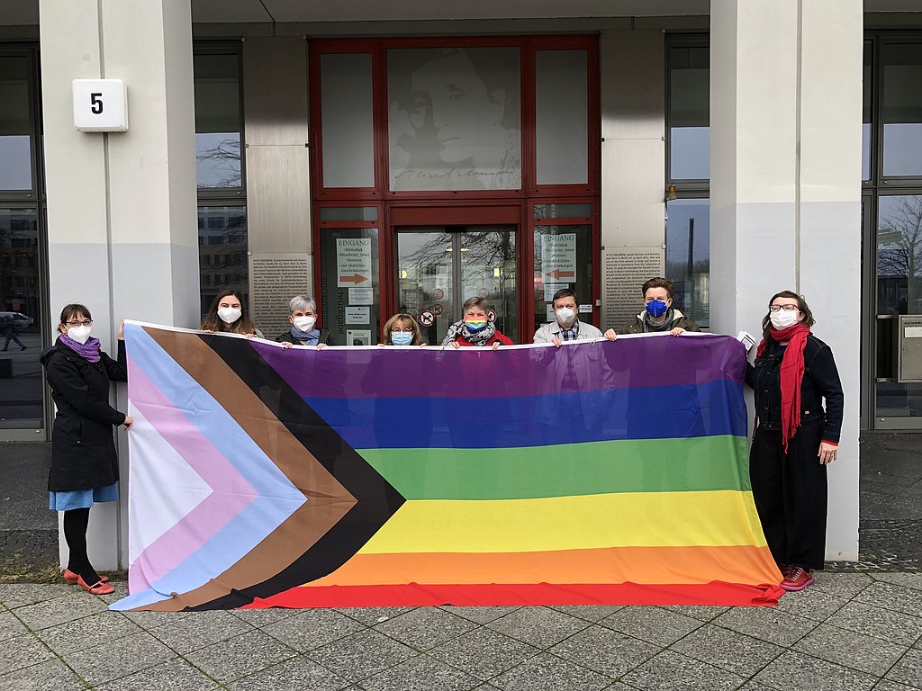 Die Kanzlerin der ASH Berlin und Mitarbeitende der Hochschule stehen vor der ASH Berlin und halten die Progressive Pride Flag hoch