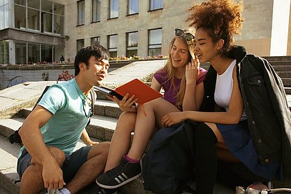 Drei Studierende unterschiedlicher Nationalität sitzen vor einem Uni-Campus in der Sonne