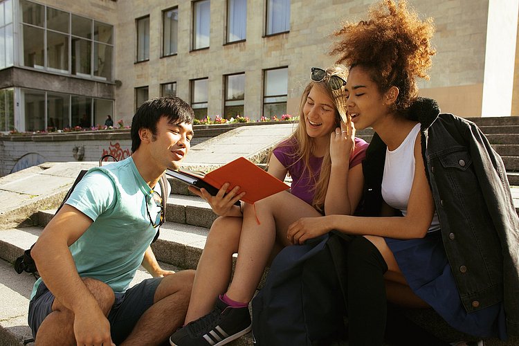 Drei Studierende unterschiedlicher Nationalität sitzen vor einem Uni-Campus in der Sonne