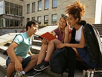 Drei Studierende unterschiedlicher Nationalität sitzen vor einem Uni-Campus in der Sonne