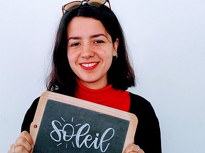 Bild der Studierenden beim Praktikum im Ausland, in Frankreich - sie hält eine Tafel mit der Aufschrift "soleil" und lächelt in die Kamera
