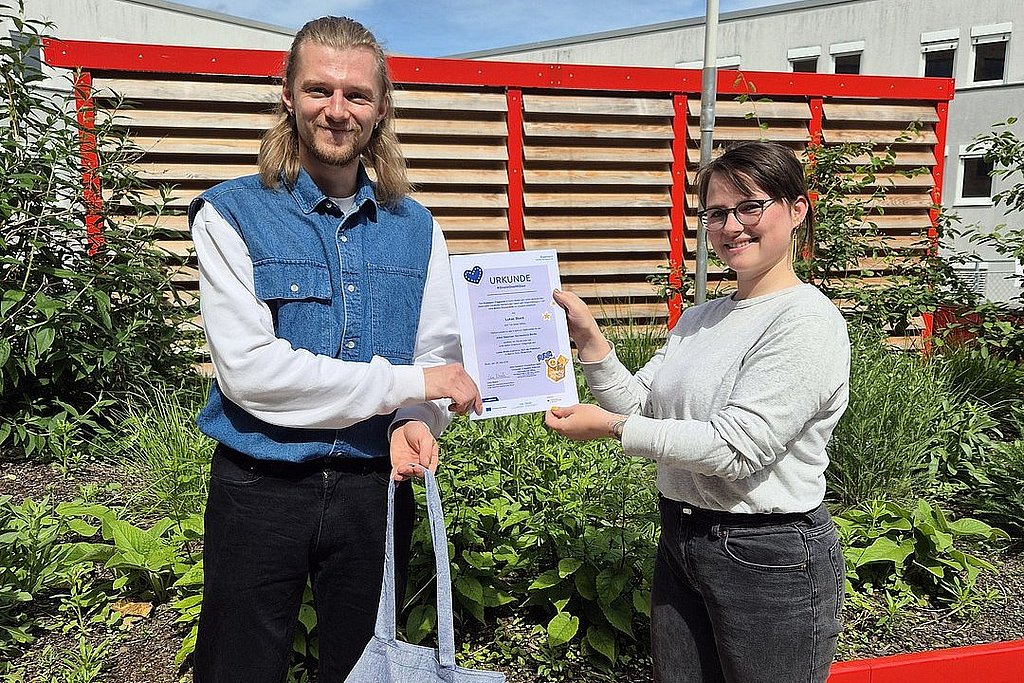 Auf dem Foto sind der ASH-Student Lukas zu sehen und eine Mitarbeiterin des International Office der ASH Berlin. Sie stehen zusammen auf der Dachterasse der Hochschule und halten eine Urkunde und einen Stoffbeutel in der Hand