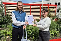 Auf dem Foto sind der ASH-Student Lukas zu sehen und eine Mitarbeiterin des International Office der ASH Berlin. Sie stehen zusammen auf der Dachterasse der Hochschule und halten eine Urkunde und einen Stoffbeutel in der Hand