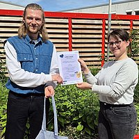 Auf dem Foto sind der ASH-Student Lukas zu sehen und eine Mitarbeiterin des International Office der ASH Berlin. Sie stehen zusammen auf der Dachterasse der Hochschule und halten eine Urkunde und einen Stoffbeutel in der Hand