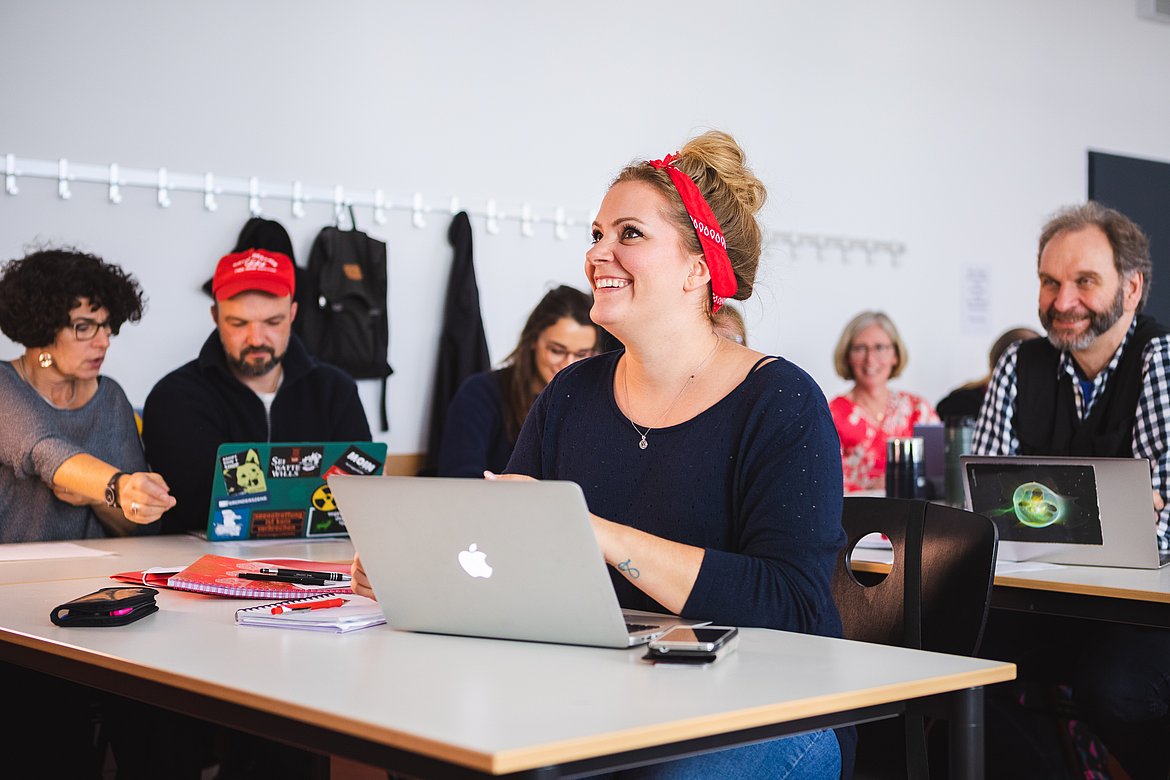 Studentin sitzt lachend vor dem Laptop im Seminarraum. Im Hintergrund eine sitzende Gruppe Studierender.