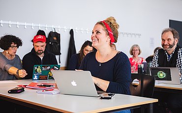 Studentin sitzt lachend vor dem Laptop im Seminarraum. Im Hintergrund eine sitzende Gruppe Studierender.